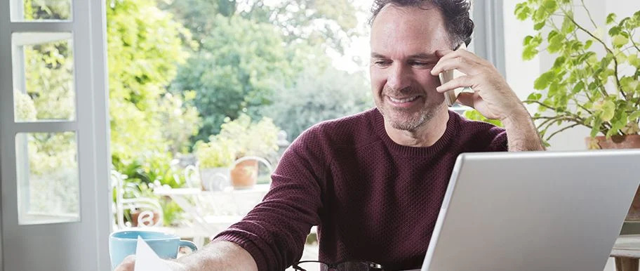 Homme souriant travaillant sur un ordinateur portable tout en passant un appel dans un espace lumineux
